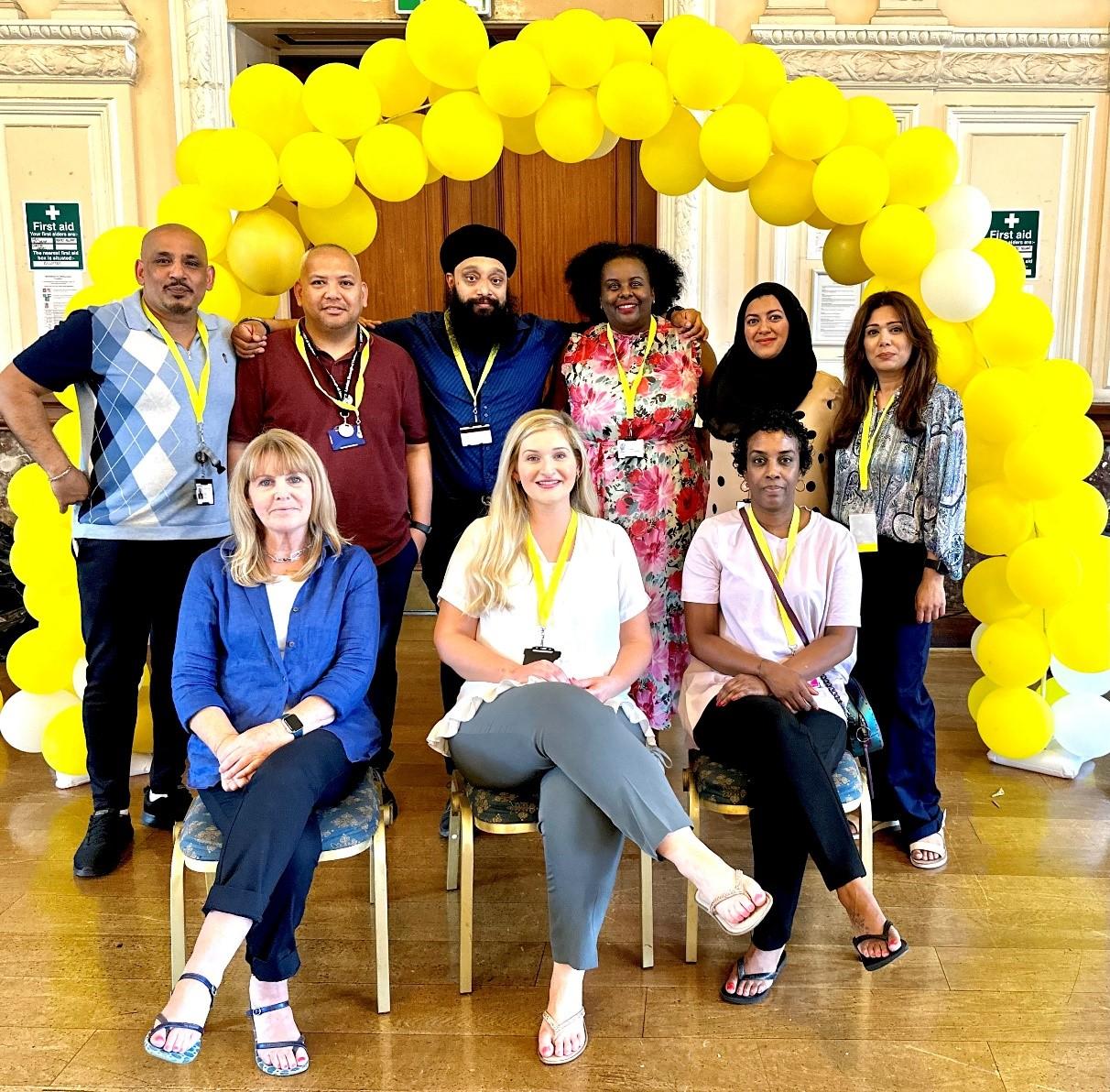 Photo of the Community Neighbourhood Link Workers team sat together in a hall in front of a yellow balloon arch